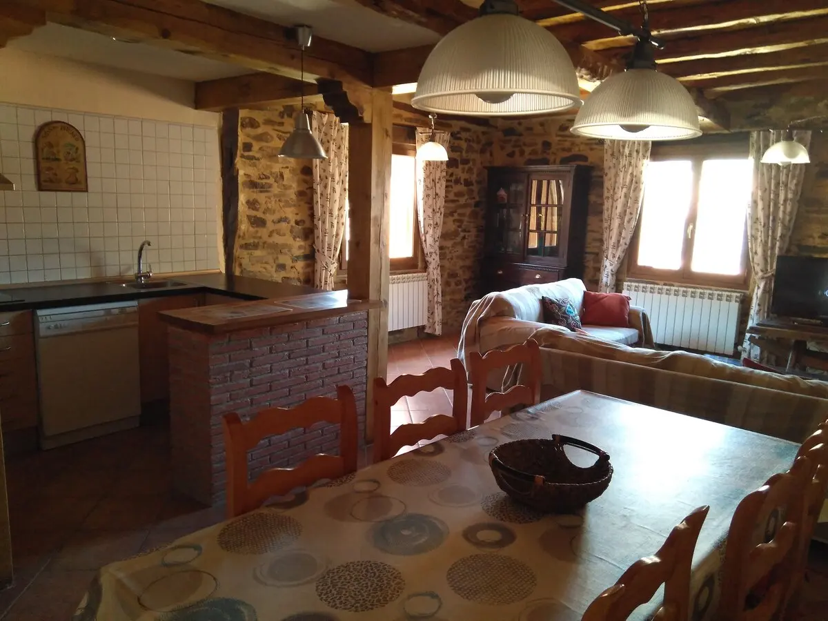 Salón y cocina en un mismo espacio, con vigas de madera, paredes de piedra y mesa de comedor